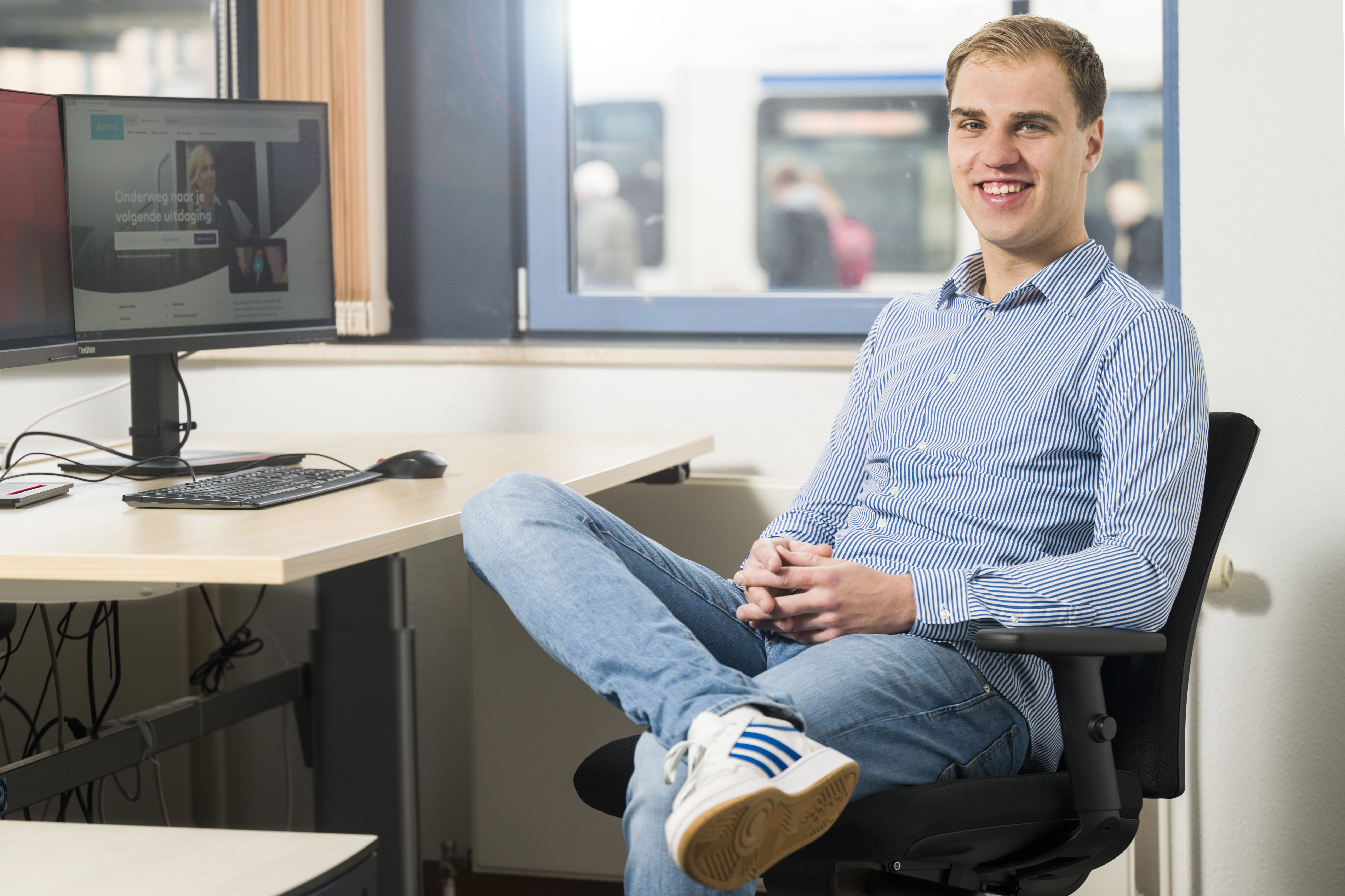 Jonge man in casual kleding zit glimlachend in bureaustoel voor een bureau met computer, achtergrond toont raamuitzicht op trein.