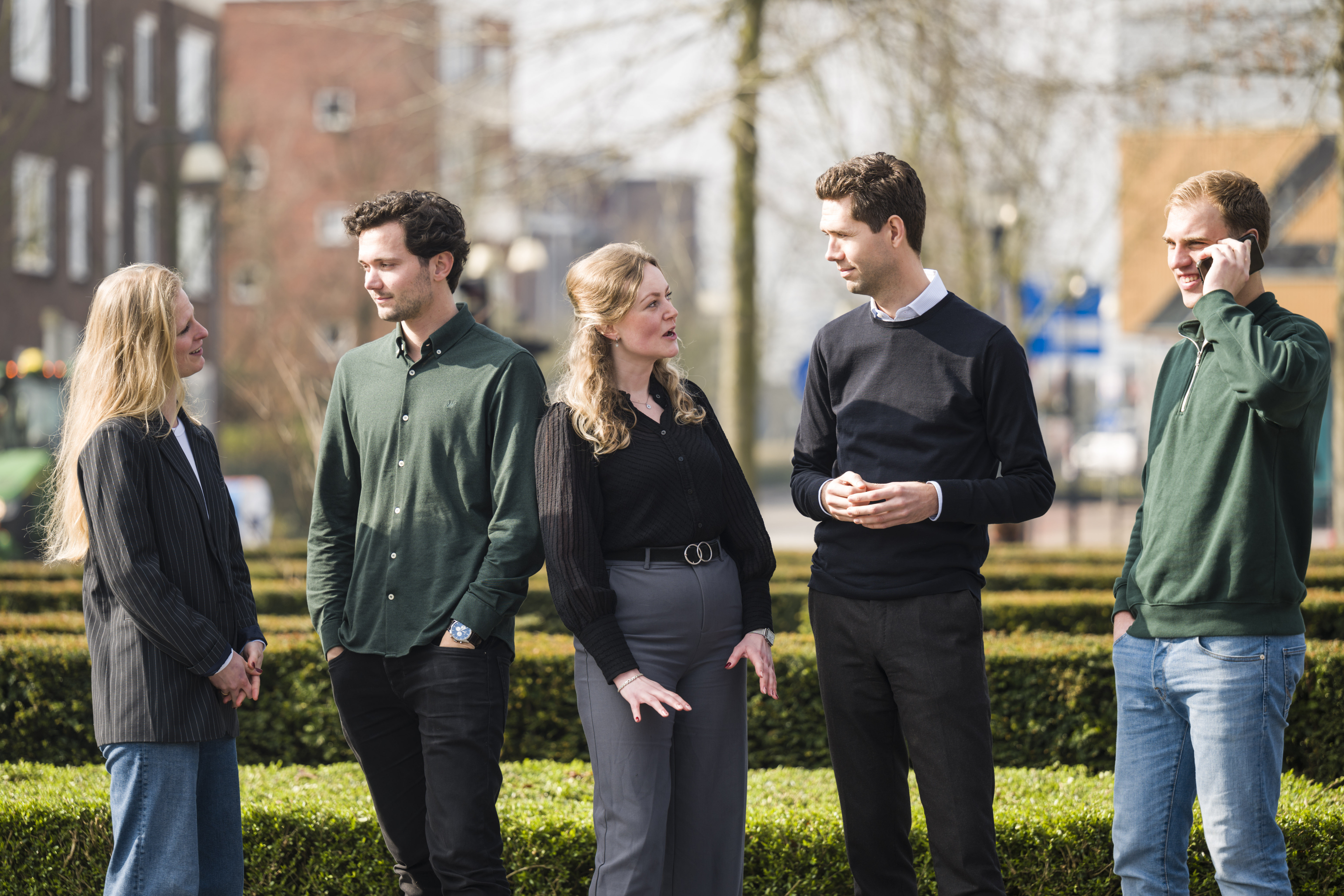 Vijf trainees in zakelijke kleding voeren levendig gesprek buiten in kring op zonnige dag.