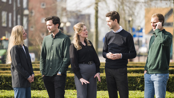 Vijf trainees in zakelijke kleding voeren levendig gesprek buiten in kring op zonnige dag. Vijf trainees in zakelijke kleding voeren levendig gesprek buiten in kring op zonnige dag.