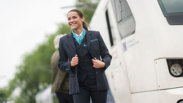 Machinist van Arriva lacht en trekt haar uniform recht, achter haar zie je een reiziger en een trein. Machinist van Arriva lacht en trekt haar uniform recht, achter haar zie je een reiziger en een trein.