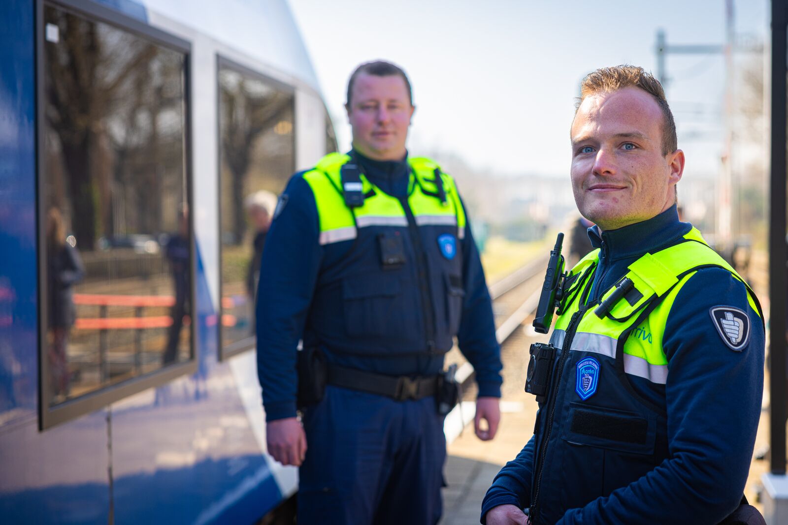 Twee veiligheidsmedewerkers in uniform staan bij een trein op een perron. Eén medewerker kijkt naar de camera, terwijl de ander op de achtergrond staat.