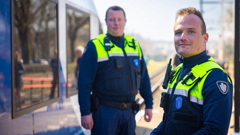 Twee veiligheidsmedewerkers in uniform staan bij een trein op een perron. Eén medewerker kijkt naar de camera, terwijl de ander op de achtergrond staat. Twee veiligheidsmedewerkers in uniform staan bij een trein op een perron. Eén medewerker kijkt naar de camera, terwijl de ander op de achtergrond staat.