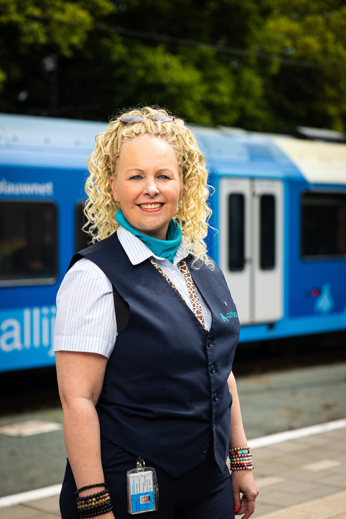Een Steward BOA-OV staat glimlachend op een perron, met een blauwe trein op de achtergrond.
