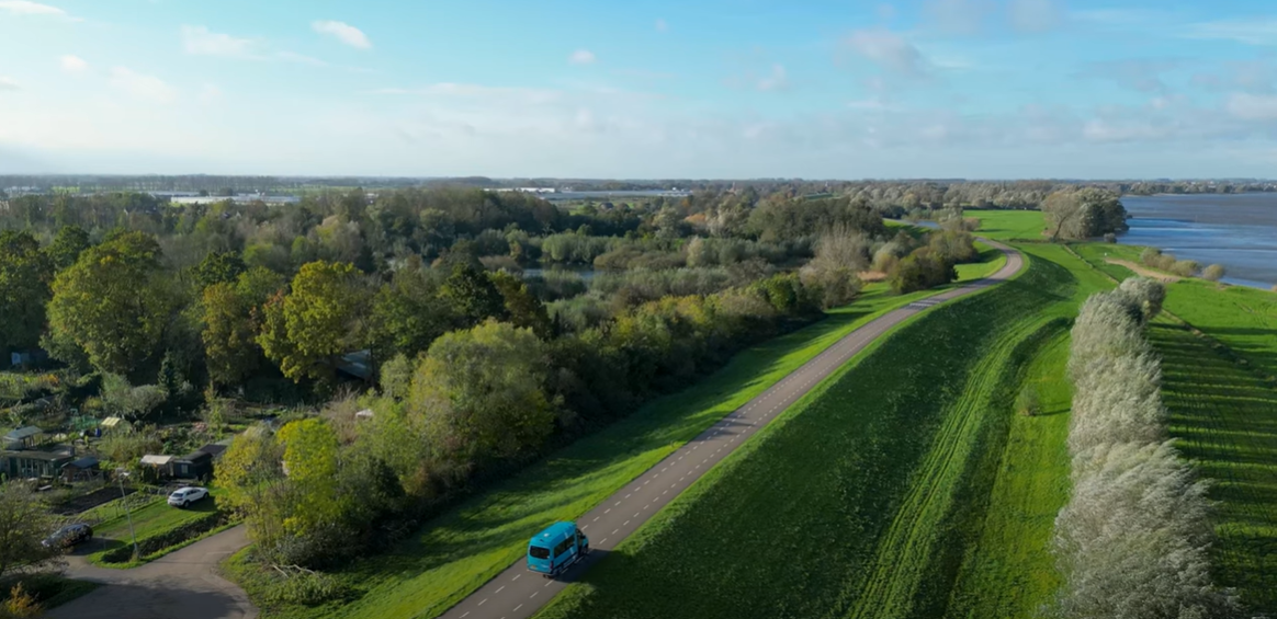 Filmstill van een bovenaanzicht van en weg op een dijk met daarop een groene Arriva-buurtbus.