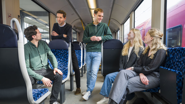 Vier personen in Arriva-trein, twee zittend, twee staand, in levendige discussie. Gekleed in smart casual, duidt op werkgerelateerde reis. Vier personen in Arriva-trein, twee zittend, twee staand, in levendige discussie. Gekleed in smart casual, duidt op werkgerelateerde reis.