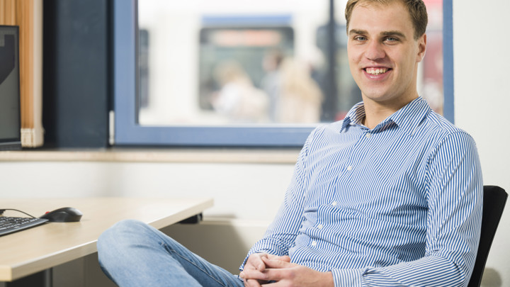 Man zit aan bureau voor computer, glimlachend, in gestreept shirt. Raam toont druk station. Man zit aan bureau voor computer, glimlachend, in gestreept shirt. Raam toont druk station.