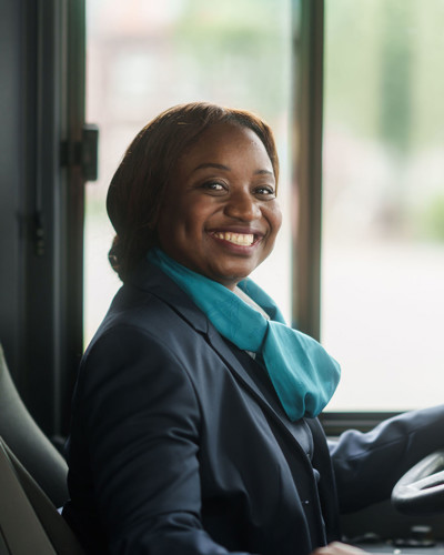 Een buschauffeur in haar Arriva-uniform zit achter het stuur en kijkt lachend opzij. Een buschauffeur in haar Arriva-uniform zit achter het stuur en kijkt lachend opzij.