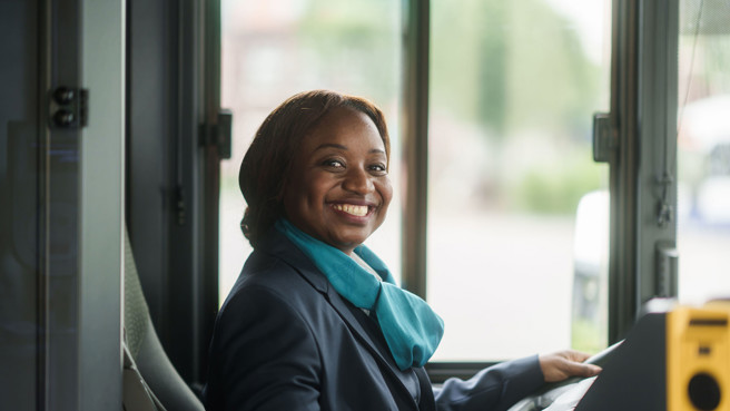 Een buschauffeur in haar Arriva-uniform zit achter het stuur en kijkt lachend opzij. Een buschauffeur in haar Arriva-uniform zit achter het stuur en kijkt lachend opzij.